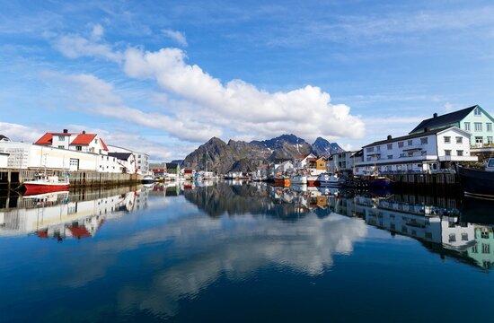 Henningsv&aelig;r harbor in Lofoten, Norway, transforms into a perfect mirror, reflecting colorful waterfront houses and the jagged V&aring;gakallen peaks under a brilliant blue sky. A breathtaking Arctic vista!