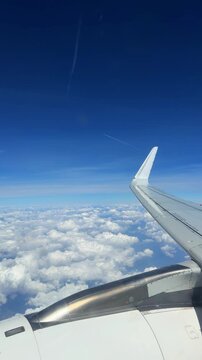 Airplane wing visible above thick white clouds under blue sky with thin contrail from other aircraft flying high during long distance trip.