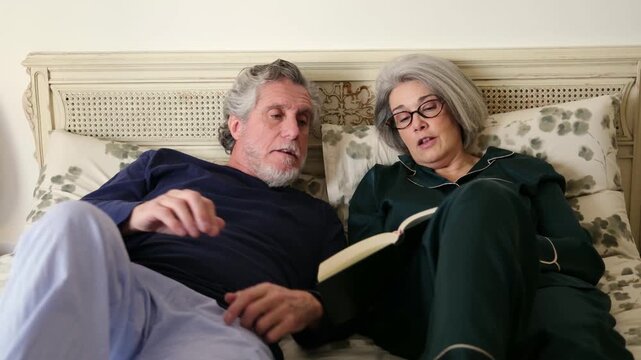 Elderly couple relaxes together, reading a book aloud in bed before sleeping