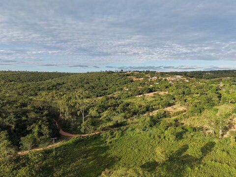Captura a&eacute;rea mostrando uma paisagem rural tranquila com pequenas propriedades, casas isoladas, vegeta&ccedil;&atilde;o nativa densa e uma estrada sinuosa sob a luz do dia.