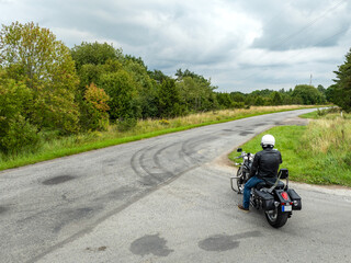 Fototapeta premium Aerial view of a lone motorcyclist standing on an empty country crossroad. The narrow road winds through a rural landscape, is flanked by fields of green bushes and trees.