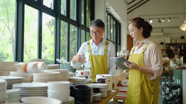 Asian male and female small business owners in yellow aprons checking inventory of handmade ceramic bowls in a bright retail pottery shop using a digital tablet for store management and sales.