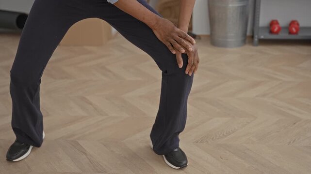 Woman touching knee in wide lunge stretch on parquet floor near rolled mats, exercise ball and rope in studio; calm focus.
