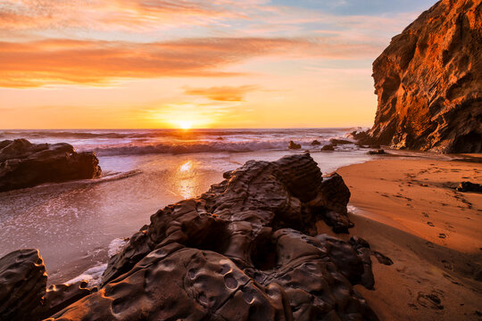 Natural sea arch at Hole in the Wall Beach in California during sunset, warm golden light illuminating rugged coastal rock, ocean waves and smooth sand, creating dramatic seaside landscape