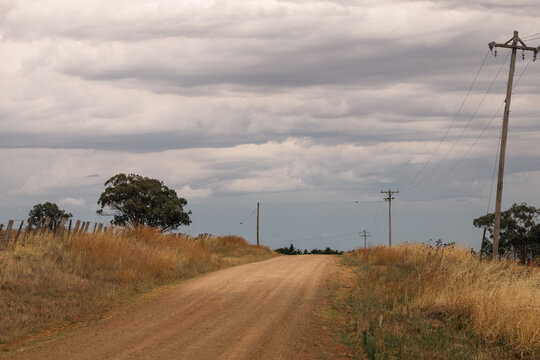 Dirt road winds through dry grass under a dramatic cloudy sky with power lines