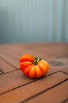 A single, ripe heirloom tomato rests on a wooden table with a blurred fence in the background