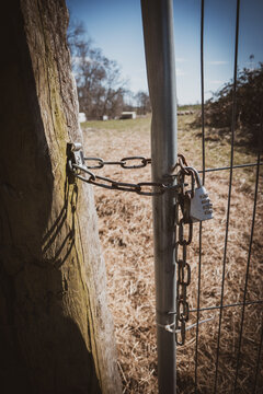 Close-up of a locked metal farm gate secured with a rusty chain and combination padlock on a sunny day in the countryside.