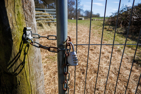 Close-up of a locked metal farm gate secured with a rusty chain and combination padlock on a sunny day in the countryside.