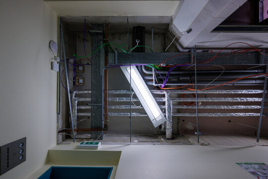 Industrial ceiling infrastructure under construction with exposed electrical wiring, cable trays, ventilation pipes, and suspended fluorescent lights in a commercial building.