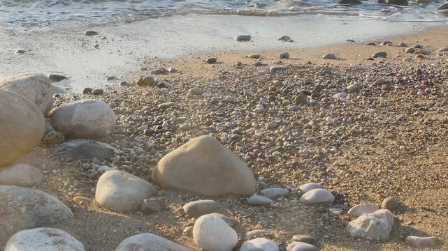 A close-up view of a pebbled beach during the golden hour, featuring gentle waves from the ocean lapping at the shore.