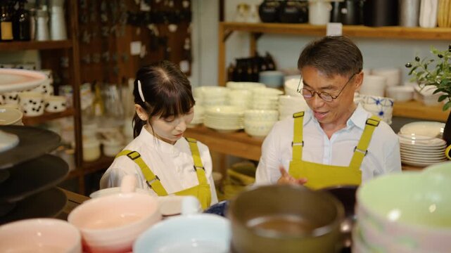 Senior Asian potter and young apprentice in yellow aprons examine ceramic bowls in a pottery studio. Experienced craftsman teaching traditional skills to a student in a ceramic shop setting.