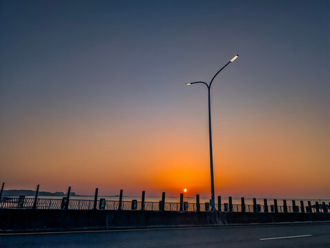 Minimalist silhouette of a double-headed street light against a vibrant orange and purple sunset sky over a calm ocean. Peaceful evening atmosphere with soft gradient colors and copy space.