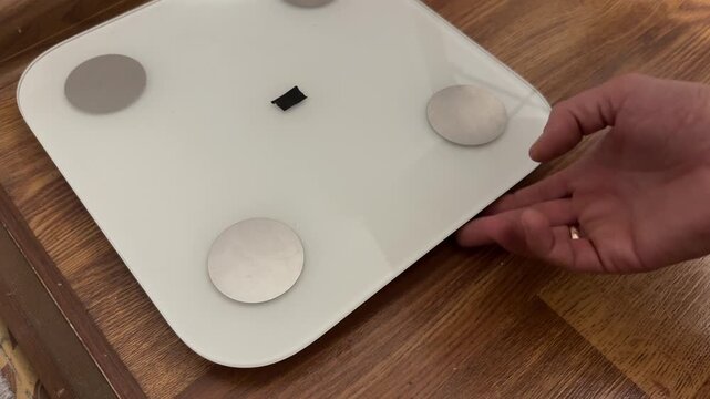 A hand touches the edge of a digital smart scale on a wooden floor