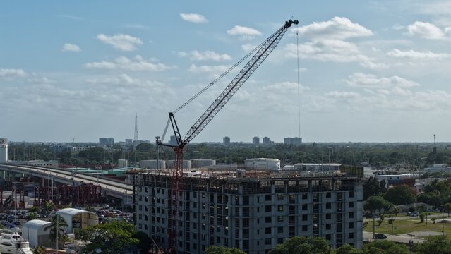 Riviera Beach, United States - 15 April 2026: Aerial view of a large construction crane atop a multi-story building under development with industrial structures and a bridge in the background.