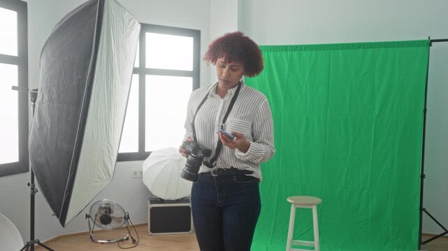 Woman photographer talks on smartphone while inspecting camera in studio with green backdrop; focused creativity.