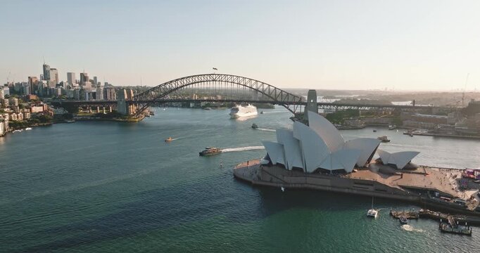 Sydney Harbour with its iconic Opera House, Harbour Bridge, and central business district skyline, boats and a cruise ship navigating the water under a clear sky. Aerial view cityscape, static shot