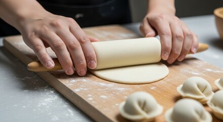 Obraz premium Close-up of hands rolling out raw white dough with a wooden rolling pin on a floured board to make homemade dumplings or pierogi, with finished dumplings in the foreground of a kitchen.