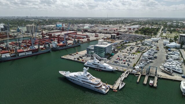 Riviera Beach, United States - 15 April 2026: Aerial view of luxury yachts docked at the marina with cargo ships and industrial port facilities in the background under a cloudy sky.