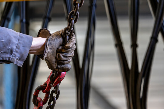 Close up of industrial worker gripping heavy steel chain with protective glove during lifting operation inside factory with crane cable system behind