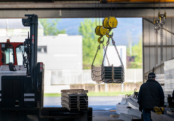 Overhead crane lifting stacked steel beams with chains inside industrial warehouse while worker walks near forklift and metal storage area © JairoMZ