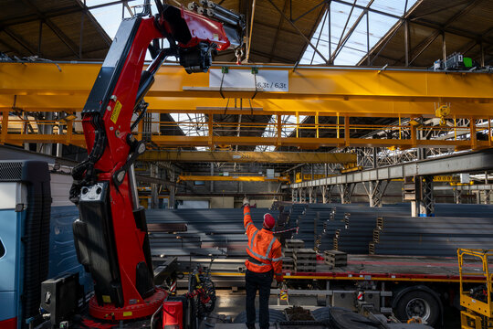 Industrial worker guiding overhead crane and red truck mounted lifting arm while transporting steel beams inside large warehouse facility