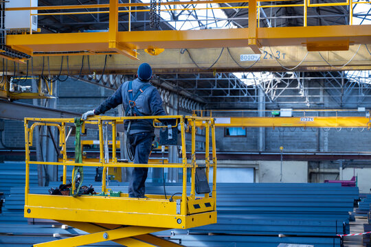 Industrial worker standing on elevated yellow scissor lift platform inside warehouse with overhead crane systems and stacked steel materials