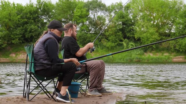 Man and woman enjoying a quiet day fishing together, sitting on folding chairs by the river with their rods in the water, relaxing during a weekend getaway in the beautiful countryside