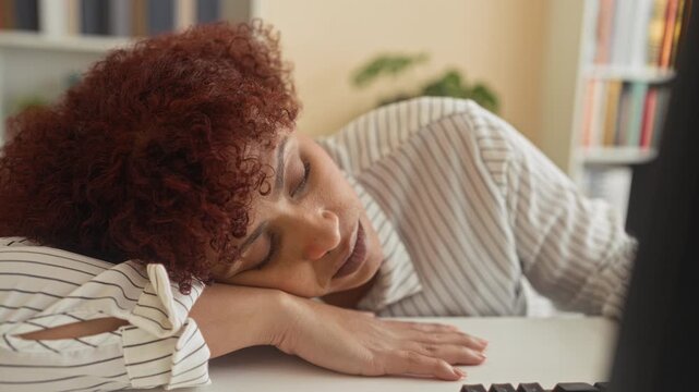 Woman resting head on desk with face on hand beside keyboard and monitor in building, striped blouse, curly red hair, bookshelves and potted plant visible; fatigue.
