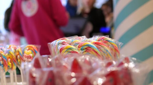 Vibrant assortment of rainbow colored candy canes and lollipops, individually wrapped in plastic, is on display for sale in a confectionery store with employees working in the background