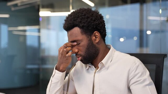 Tired african american businessman suffering from eye strain and headache while sitting at workplace in business office. Male worker takes off glasses and massages, rubs bridge of nose. Close up