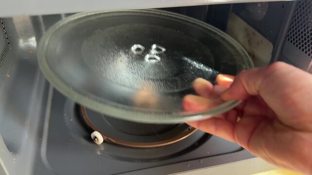 A person carefully removes a round glass plate from a microwave oven. The gesture suggests cleaning, repair, or rearranging