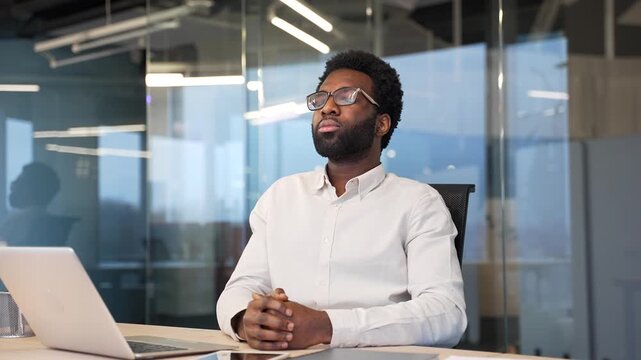 Calm african american businessman resting with his eyes closed breathing deeply while sitting at desk at workplace in a modern business office. Happy worker relaxing in break at work, relieves stress