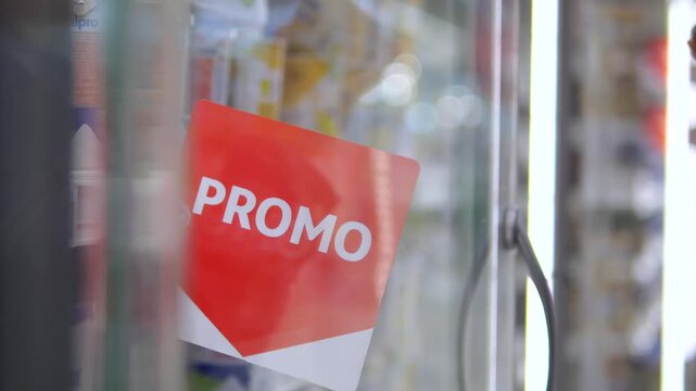 Close up on a red promotional sign as a customer's hand opens a refrigerator door in a grocery store to choose a discounted product from the shelf, highlighting sales and consumerism