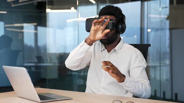 African american works using VR glasses in virtual reality simulator sitting at workplace in modern business office. Worker uses gestures to control computer programs and flips through virtual pages