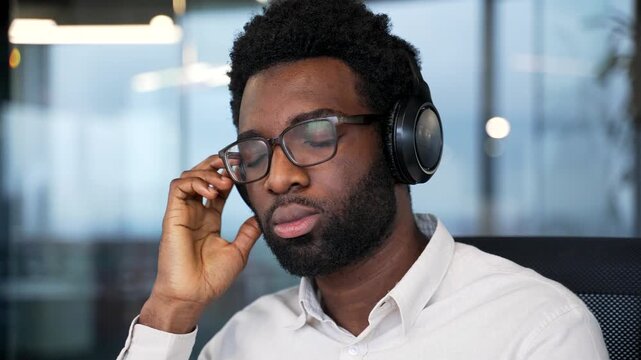 African american businessman in headphones listening to music with closed eyes sitting at workplace in a business office. Happy worker relaxes to sounds of relaxation and noise of nature. Close up