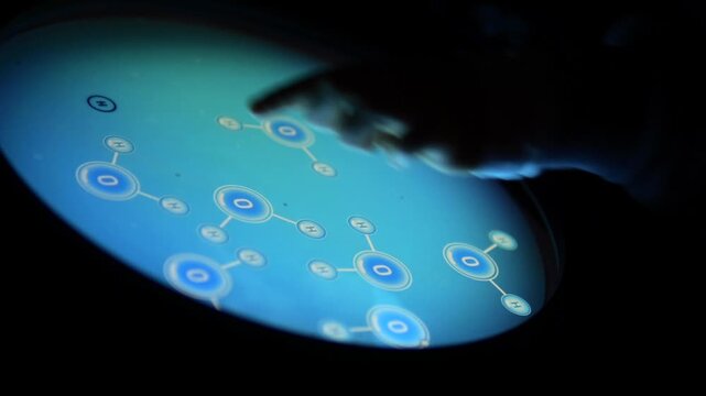 Young student's hand touching an educational, interactive screen showing water molecules, learning chemistry in a dark, modern science museum environment with a futuristic, glowing display