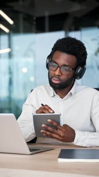 African american businessman in headphones watching video call conference takes notes looking at laptop screen sitting at workplace in business office. Worker listening online training. Vertical video