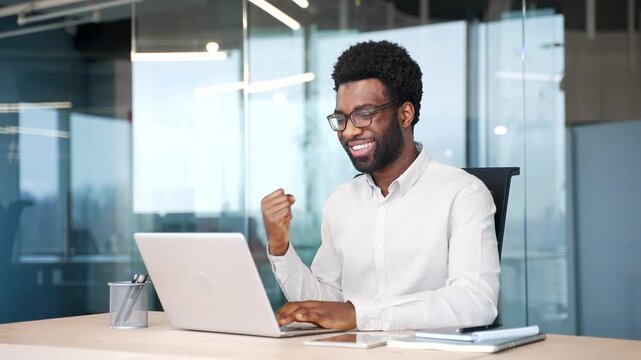 Satisfied happy african american businessman received great news on laptop sitting at desk at workplace in modern business office. Smiling glad male worker reads positive good message on computer