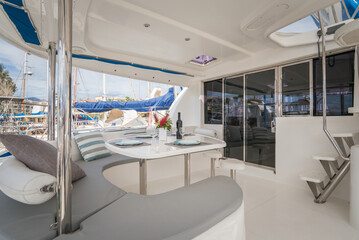 Obraz premium Overview of exterior lounge of a modern charter sailing catamaran, with cushions and dining table set, flowers and bottle of wine, located in a marina in Caribbean
