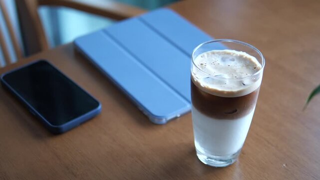 Cinematic close-up of layered iced coffee latte on a wooden table with smartphone and tablet, modern remote work and lifestyle concept