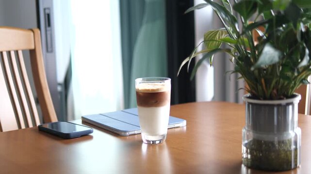 Cinematic wide shot of a layered iced coffee latte on a wooden desk with a green plant, tablet, and smartphone in a bright interior