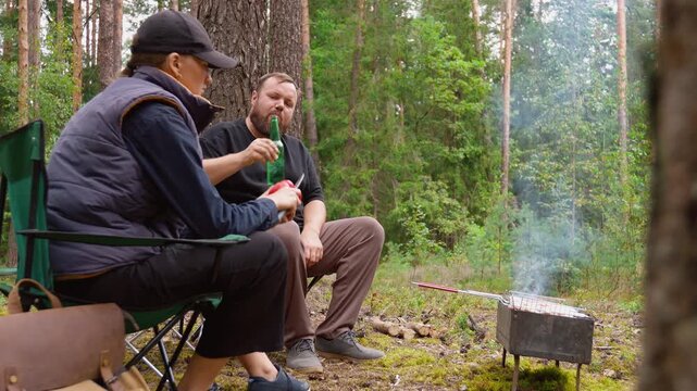 Bearded man cooking meat on a charcoal grill while his partner relaxes in a chair during a peaceful picnic in a pine forest, enjoying nature and outdoor recreation together