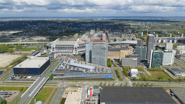 Amsterdam, Netherlands - 19 April 2026: Aerial view of the Johan Cruijff Arena stadium surrounded by modern office buildings and urban infrastructure under a cloudy sky.