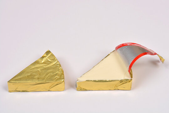 A side view of two triangular processed cheese portions wrapped in gold foil with one being partially opened to reveal the creamy cheese inside