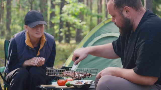 Young man and woman enjoying picnic in beautiful forest, bearded man grilling meat on barbecue with tent and laying it out on table with grill grate