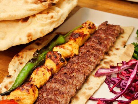 A vibrant close-up of an authentic Middle Eastern mixed grill platter. The dish features Adana kebab, chicken shish, and a grilled green chili on flatbread, accompanied by roasted tomatoes, pickled