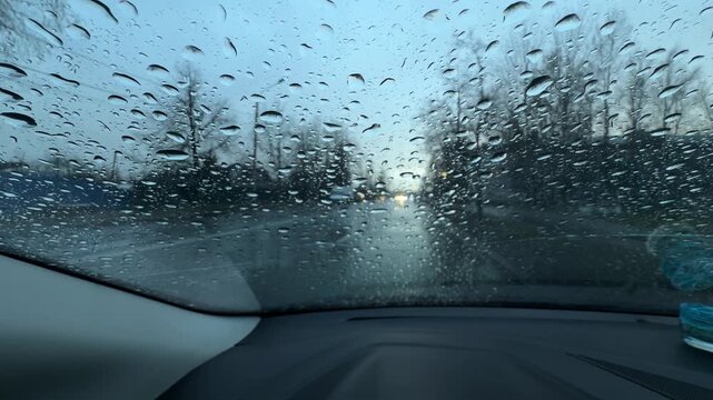 View through a wet windshield with raindrops while driving on a city road. Cars, bare trees, and an overcast sky are visible ahead.