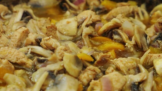 Chicken, onions, and champignons are fried in vegetable oil in a deep cast-iron skillet. Medium shot.