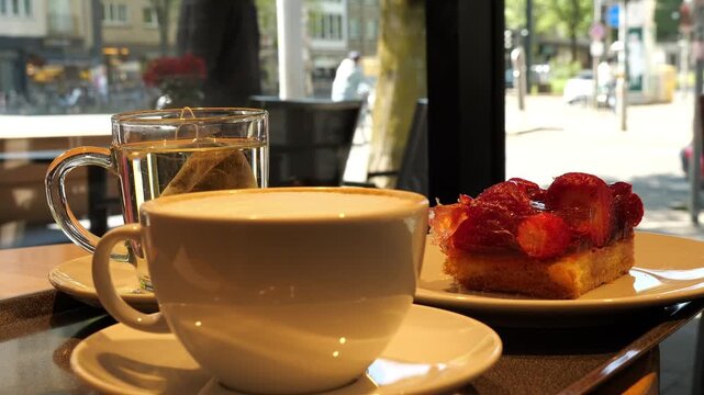 A cozy cafe scene featuring a clear glass mug with a tea bag steeping in hot water and a white ceramic cup of cappuccino on a tray. A strawberry tart is partially visible. The setting is by a window, 