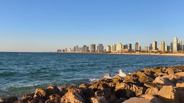 Dynamic daytime footage of a deep blue, turbulent sea with waves in constant motion. In the distance, a modern city skyline with high-rise buildings, a rugged shoreline covered with numerous stones